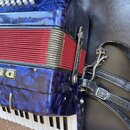 Detailní pohled na modrou akordeon s perleťovým povrchem a červeným méchem. Na instrumentu jsou vidět bílé klávesy a knoflíky, k tělu jsou připevněny…