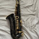 Černý saxofon s pozlacenými klapy a mechanikou. Na korbide je vyryt ornament s květinami. Nástroj nemá žádné viditelné značky ani nápisy. Povrch je g…