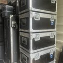 Snímek zobrazuje stoh pěti černých transportních case s hliníkovými hranami a kovovým kováním. Na některých kufrech je vidět modrý štítek s bílým náp…