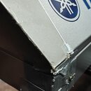 Detail levého horního rohu stříbřitého klávesového nástroje s modrým logem a nápisem YAMAHA. Roh je výrazně poškozený, materiál je odloupaný a v míst…