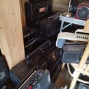 Fotografie zachycuje hromadu starších černých audio systémů a přenosných radiokasetových přehrávačů v dřevěném prostoru. Na jednom z přístrojů je vid…