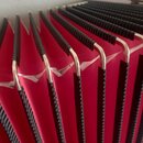 Detailní pohled na červené měchy akordeonu s černými hranami. V horní části jsou vidět kovové lišty, které vykazují známky opotřebení a roztržení čer…