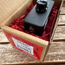 Černý efekt pedál v kartonové krabici s červenou výplní. Na přední straně krabice je náleplka s nápisy LITTLE BLACK AMP BOX, SERIAL a JHS PEDALS spol…