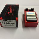 Červený efekt pedál s nápisem Maxon CP9Pro+ Compressor/Limiter a stříbrným kovovým stupadlem. Vedle pedálu je černá krabice s červenými prvky a nápis…