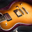 Elektrická kytara s tmelem v odstínu honey burst a bílým lemováním korpusu leží v černém kufru s fialovou výstuhou. Nástroj má dva humbuckery, dva ov…