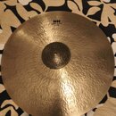 Zlatavý kovový činel s viditelným kovánívým povrchem a klepaným středem. V horní části je černý nápis HH, pod ním text Rare Bell Dry Ride a dále 21"/…