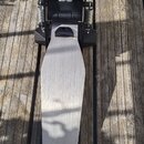 Na dřevěném podkladu leží elektronický pedál basového bubnu v černé a stříbrné barvě. Na spodní části stříbrného šlapadla je umístěn nápis Roland. V…