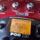 Detailní pohled na červený procesor Line 6 POD X3. Na horní části jsou stříbrné kovové ovratidla s nápisy MIDDLE, TREBLE, PRESENCE a REVERB. Uprostře…