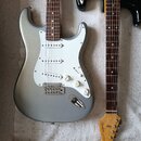 Stříbrná elektrická kytara typu Stratocaster s bílým pickguardem a hnědým hmatníkem. Na kytare jsou tři bílé oválné knofíky a chromovaný most s pruži…