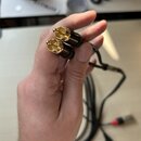Ruka drží dva zlaté konektory typu RCA s černým plastovým tělem. K konektorům vede černý kabel, který je v pozadí rozvinutý a zakončen dalším konekto…