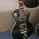 Černá elektrická kytara typu Les Paul s bílým lemováním. Na těle jsou dva humbuckery, přibyvková kobylka a jedna ovládacív čára. V dolní části těla j…