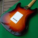 Zadní strana elektrické kytary s tvarem typu Stratocaster v barevném přechodu sunburst (černá, červená a žlutá). Uprostřed těla je bílý plastový kryt…