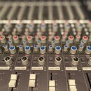 Detailní pohled na ovládací panel míchací konzole s řadami potenciometrů a posuvníků. Na panelu jsou vidět nápisy DAW, SOURCE, PAN, SHORT FADER a MIX…