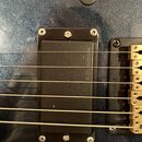 Detailní záběr na černý humbucker s krytem, který je přišroubován k tmavě modrému třpytivému povrchu elektrokitary. Na pravé straně je vidět část zla…