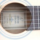Detailní pohled na ozvučnici akustické kytary světlého dřeva s nápisem vpravo od hmatníku. Uvnitř ozvučnice je štítek s nápisy ORTIGA GUITARS • 1ST 1…
