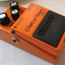 Oranžový efekt pedál s černým plastovým spínacím tlačítkem. Na horní straně je nápis Distortion DS-1 a logo BOSS, tři černé ovládací knoflíky s popis…
