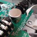 Detailní pohled na zelenou základní desku s elektronickými součástkami, včetně velkého kovového kondenzátoru a černého chladiče. Na desce jsou bílé n…