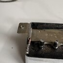 Detailní pohled na kovovou součástku, pravděpodobně vnitřek efektového pedálu nebo elektroniky nástroje. Na kovovém povrchu je vyraženo číslo 7,842 a…