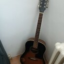 Akustická kytara s tmavým korpusem v provedení sunburst. Na hlavě hmatníku je bílý nápis Epiphone. Nástroj je opřen o bílou zeď vedle radiátoru.