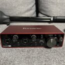 Červené audio rozhraní značky Focusrite, model Scarlett 2i2. Přední panel obsahuje dva vstupy s přepínači INST a AIR, tlačítko 48V, funkci DIRECT MON…
