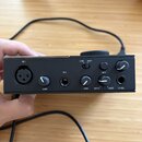 Černé audio rozhraní s kovovým povrchem. Na předním panelu jsou nápisy IN 1, IN 2, LINE, INST, GAIN, INPUT, HOST a VOL, dále přepínač pro 48 V. Zaříz…