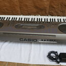 Šedý elektronický klavír značky CASIO s bílými a černými klávesami. Na přední straně je velký nápis CASIO a vedle něj čtyři konektory. Před nástrojem…
