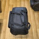Černá textilní přepravní taška s rukojetí a dvěma popruhy. Taška je umístěna na dřevěné podlaze. Povrch materiálu vykazuje mírné znečištění a opotřeb…