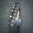 Černý hlavník elektrické kytary s bílým nápisem Ibanez. Na horní hraně je drobný nápis RG SERIES. Jsou vidět kovové ladičky a struny, povrch je gloss…