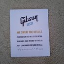 Fotografie znázorňuje bílou papírovou kartu s textem. V horní části je tmavě modré logo Gibson a pod ním nápis usa. Uprostřed je zlatý text WE SWEAT…