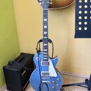 Modrá elektrická kytara typu Les Paul s dvěma humbuckery a bílým pickguardem, umístěná na černém stojanu. Na hlavě hmatníku je nápis Gibson a modelov…
