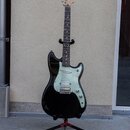 Černá elektrická kytara s bílým pickguardem umístěná na černém stojanu. Na hlavě hmatníku je viditelné logo Fender. Nástroj má tři snímače typu singl…