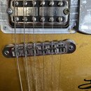 Detail elektrické kytary v zlaté barvě s kovovým snímačem a kobylkou. Na krytu snímače je vyražen nápis U.S.PAT 2892471. Povrch kovu vykazuje známky…