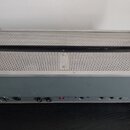 Šedý elektronický přístroj s kovovým perforovaným krytem v horní části. Na spodním zadním panelu jsou umístěny přepínače, konektory a ovládací prvky.…