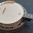 Bílé banjo s chromovaným kroužkem a hradítkem, položené na modrém koberci. Na membráně je vidět logo znázorňující korunu a nápis „MADE IN USA BY COBB…