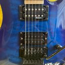Detail elektrické kytary v modré barvě s výraznou strukturou dřeva. Nástroj má dva černý humbuckery, přičemž na horním je bílý nápis Jackson. V dolní…
