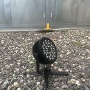 Černý LED reflektor s kulatou hlavou a 12 svítícími elementy v mřížce. Zařízení je umístěno na černém kovovém stativu s nohama. Povrch těla je žebrov…
