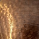 Detailní záběr na povrch kovového činele v měděné barvě s viditelnou strukturou kování. Na povrchu je vyryto logo a nápis PAISTE, pod kterým je styli…