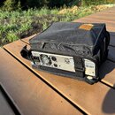 Černý textilní přepravní pouzdro s oranžovým logem na horní části, ve kterém je umístěno stříbrné elektronické zařízení. Na zadní straně přístroje js…