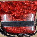 Červená akordeon s marbled povrchem a černým vrškem. Na instrumentu je viditelný nápis Made in Germany a další méně čitelné znaky. Nástroj má připevn…