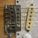 Detail elektrické kytary s chromovaným mostem a bílým pickguardem s perleťovým vzorem. Na krémovém snímači je viditelný nápis "Music Man". Povrch mos…
