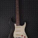 Černá elektrická kytara typu Stratocaster s bílým pickguardem a tři červenými oválnými knoflíky. Hmatník a hlava jsou vyrobeny z dřeva, na hlavě je v…