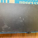 Detailní pohled na černý, texturovaný povrch hudebního zařízení. Povrch je viditelně opotřebovaný, s četnými škrábanci a odřeninami. V horní části je…