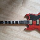 Červená elektrická kytara typu SG s tmavým hmatníkem a bílými vložkami. Na těle jsou dva humbuckery, čtyři oválné knofíky a přepínač. Na pravé straně…