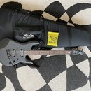 Šedá elektrická kytara s matným povrchem, černým hardwarem a hnědým hmatníkem. Na hlavě kytary je bílý nápis Sterling by Music Man. Kytara leží na če…