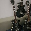 Fotografie zobrazuje tři černé elektrické kytary s tvarem Les Paul. Přední kytara má na hlavě nápis Xamanid a bílé vložky na hmatníku. V pozadí je vi…