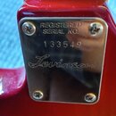 Detail kovového štítku na červeném povrchu hudebního nástroje. Na štítku je vyrazen text REGISTERED SERIAL NO. a pod ním číslo 133549, v dolní části…