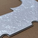 Bílý plechový kryt (pickguard) s perleťovým efektem a černým lemováním okrajů. Předmět má specifický vlnitý tvar s otvorem pro šroubek v horní části.…