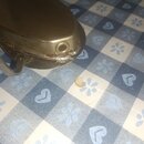 Detailní záběr na kovový, stříbrný nebo zlatavý objekt s oblým tvarem. Povrch vykazuje známky opotřebení, znečištění a v dolní části je vidět hrubé s…
