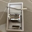 Bílá papírová krabice obsahuje kovový tremolo systém pro elektrickou kytaru, dvě pružiny a kovovou zadní desku s vruty. Součástí je také kovová páka…