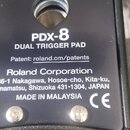 Detailní pohled na černý plastový kryt elektronického padu. Je na něm bílý nápis Roland, model PDX-8 a označení DUAL TRIGGER PAD. Dále je uveden text…